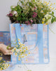 A square canvas tote bag with a pastel patchwork pattern featuring seashells, flowers, and palm trees in lavender, orange, and light blue, with solid light blue handles.  Lizzie Print - Daily Bag - Reusable bags online | Weekender bags | Shopper bags | Pouches  Hello Weekend Beige canvas handles | Beach Bags