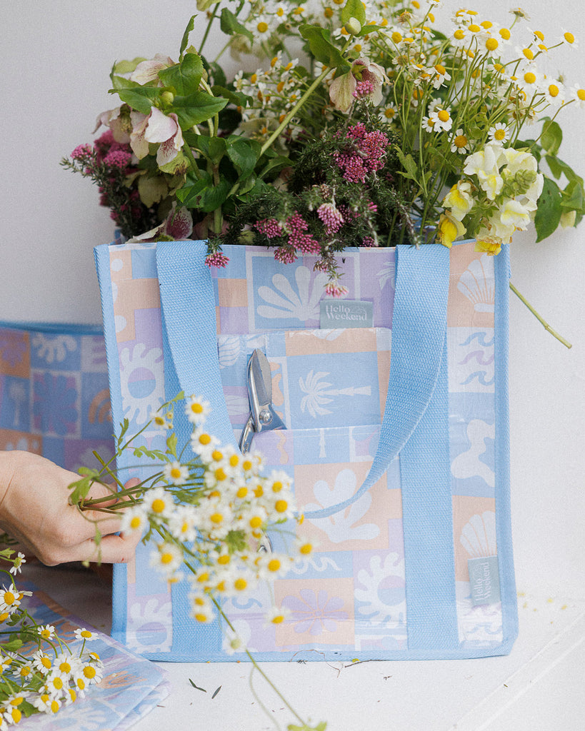 A square canvas tote bag with a pastel patchwork pattern featuring seashells, flowers, and palm trees in lavender, orange, and light blue, with solid light blue handles.  Lizzie Print - Daily Bag - Reusable bags online | Weekender bags | Shopper bags | Pouches  Hello Weekend Beige canvas handles | Beach Bags