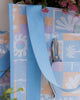 A square canvas tote bag with a pastel patchwork pattern featuring seashells, flowers, and palm trees in lavender, orange, and light blue, with solid light blue handles.  Lizzie Print - Daily Bag - Reusable bags online | Weekender bags | Shopper bags | Pouches  Hello Weekend Beige canvas handles | Beach Bags