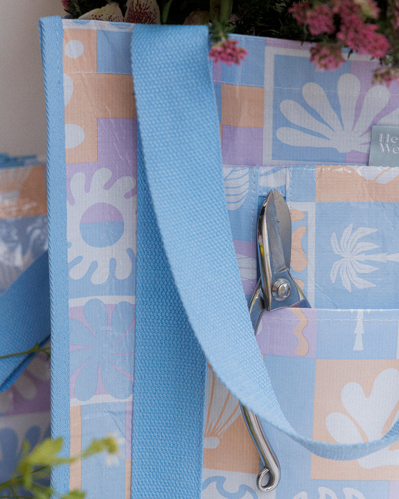 A square canvas tote bag with a pastel patchwork pattern featuring seashells, flowers, and palm trees in lavender, orange, and light blue, with solid light blue handles.  Lizzie Print - Daily Bag - Reusable bags online | Weekender bags | Shopper bags | Pouches  Hello Weekend Beige canvas handles | Beach Bags