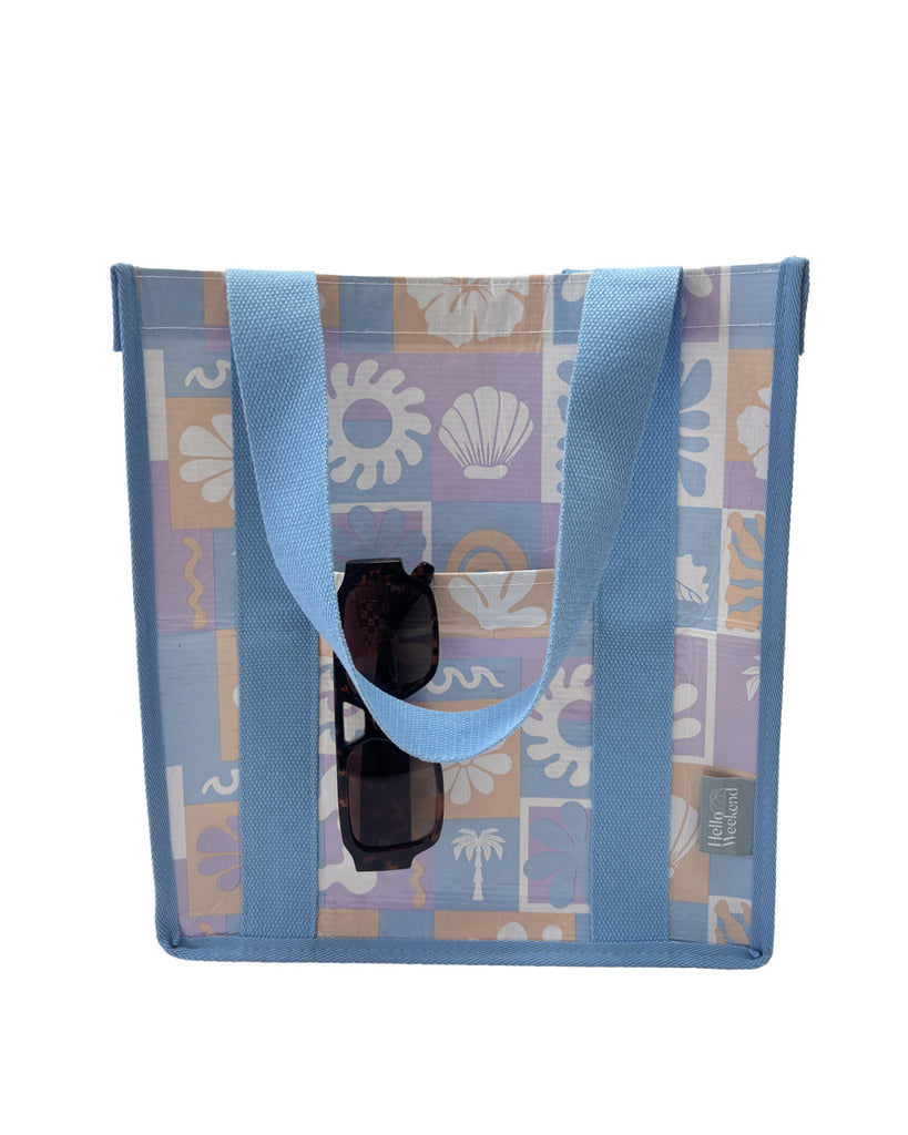 A square canvas tote bag with a pastel patchwork pattern featuring seashells, flowers, and palm trees in lavender, orange, and light blue, with solid light blue handles.  Lizzie Print - Daily Bag - Reusable bags online | Weekender bags | Shopper bags | Pouches  Hello Weekend Beige canvas handles | Beach Bags