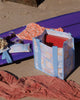 A square canvas tote bag with a pastel patchwork pattern featuring seashells, flowers, and palm trees in lavender, orange, and light blue, with solid light blue handles.  Lizzie Print - Daily Bag - Reusable bags online | Weekender bags | Shopper bags | Pouches  Hello Weekend Beige canvas handles | Beach Bags