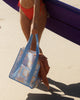 A square canvas tote bag with a pastel patchwork pattern featuring seashells, flowers, and palm trees in lavender, orange, and light blue, with solid light blue handles.  Lizzie Print - Daily Bag - Reusable bags online | Weekender bags | Shopper bags | Pouches  Hello Weekend Beige canvas handles | Beach Bags