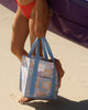 A square canvas tote bag with a pastel patchwork pattern featuring seashells, flowers, and palm trees in lavender, orange, and light blue, with solid light blue handles.  Lizzie Print - Daily Bag - Reusable bags online | Weekender bags | Shopper bags | Pouches  Hello Weekend Beige canvas handles | Beach Bags