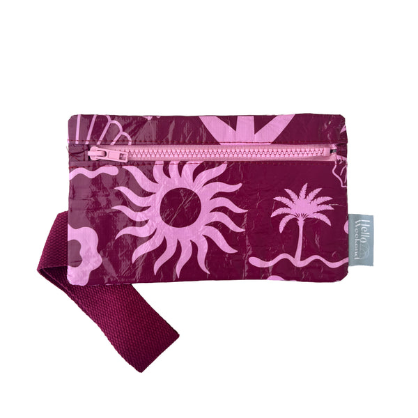A small zippered pouch with a maroon wrist strap, featuring a palm tree and sun mixed pattern of bright pink shapes on a maroon colour. A small fabric tag on the right reads Hello Weekend. Layla Print - Phone Pouch - Reusable bags online | Daily bags | Shopper bags | Weekender bags  Hello Weekend