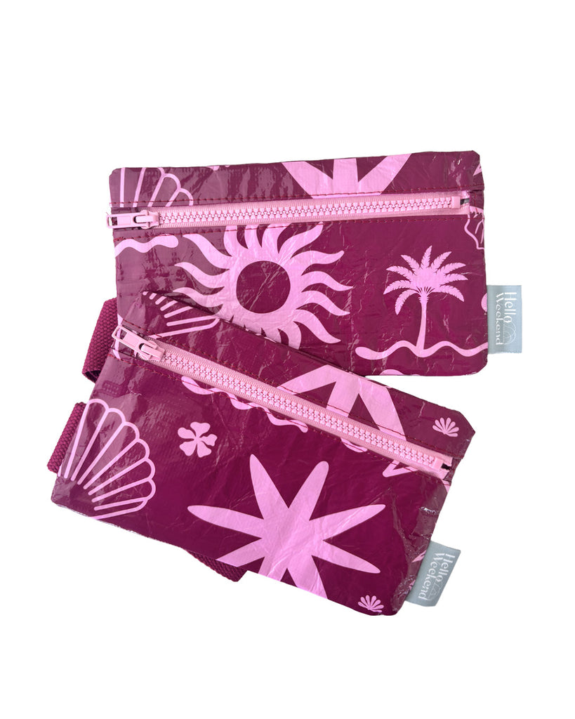 A small zippered pouch with a maroon wrist strap, featuring a palm tree and sun mixed pattern of bright pink shapes on a maroon colour. A small fabric tag on the right reads Hello Weekend. Layla Print - Phone Pouch - Reusable bags online | Daily bags | Shopper bags | Weekender bags  Hello Weekend