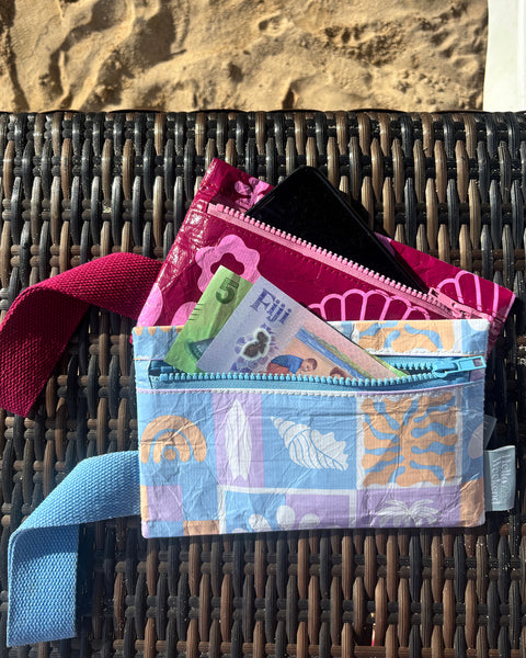 A small zippered pouch with a maroon wrist strap, featuring a palm tree and sun mixed pattern of bright pink shapes on a maroon colour. A small fabric tag on the right reads Hello Weekend. Layla Print - Phone Pouch - Reusable bags online | Daily bags | Shopper bags | Weekender bags  Hello Weekend
