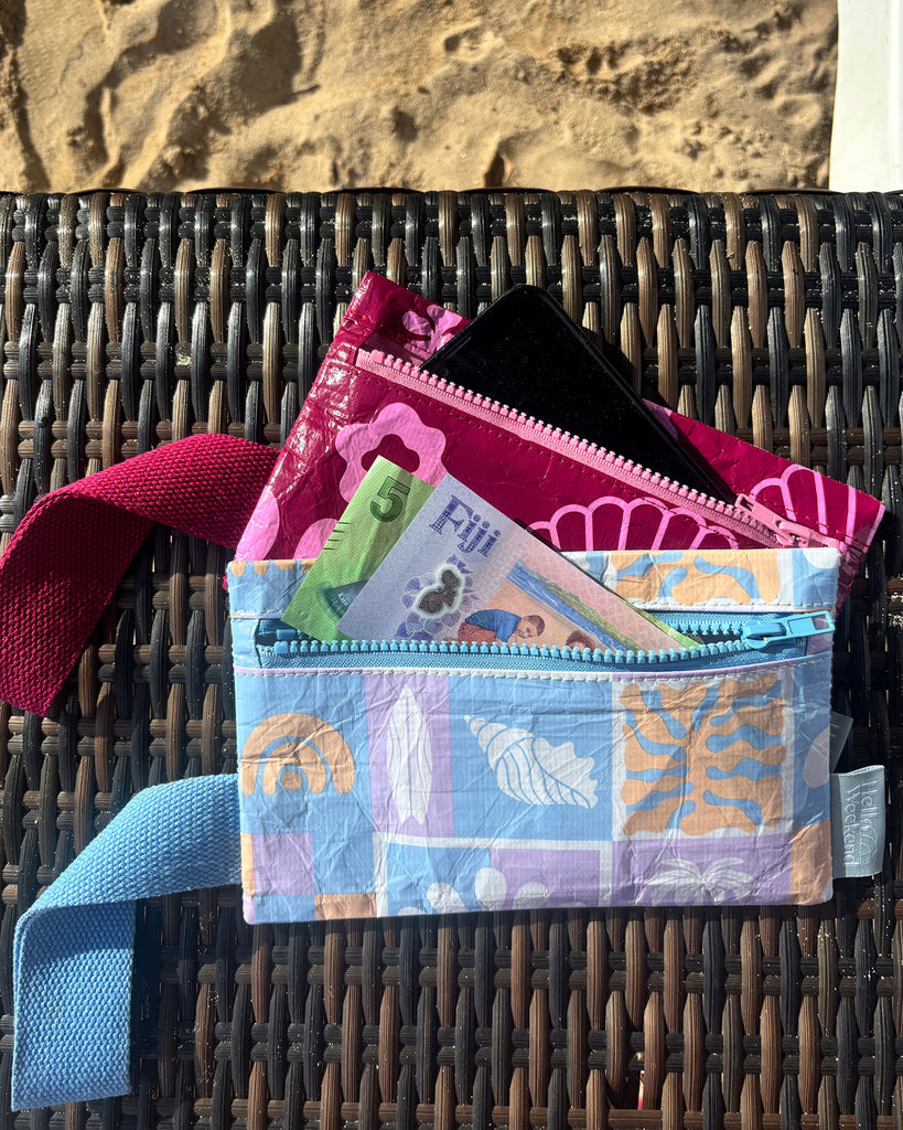 A small zippered pouch with a maroon wrist strap, featuring a palm tree and sun mixed pattern of bright pink shapes on a maroon colour. A small fabric tag on the right reads Hello Weekend. Layla Print - Phone Pouch - Reusable bags online | Daily bags | Shopper bags | Weekender bags  Hello Weekend