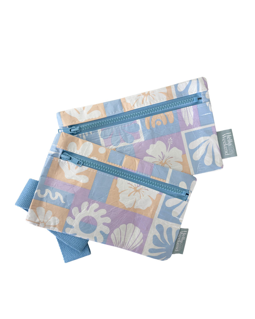 A small zippered pouch with a blue wrist strap, featuring a patchwork pattern of light blue, lavender, and peach squares with white floral and leaf motifs. A small fabric tag on the right reads Hello Weekend. Lizzie  Print - Phone Pouch - Reusable bags online | Daily bags | Shopper bags | Weekender bags  Hello Weekend