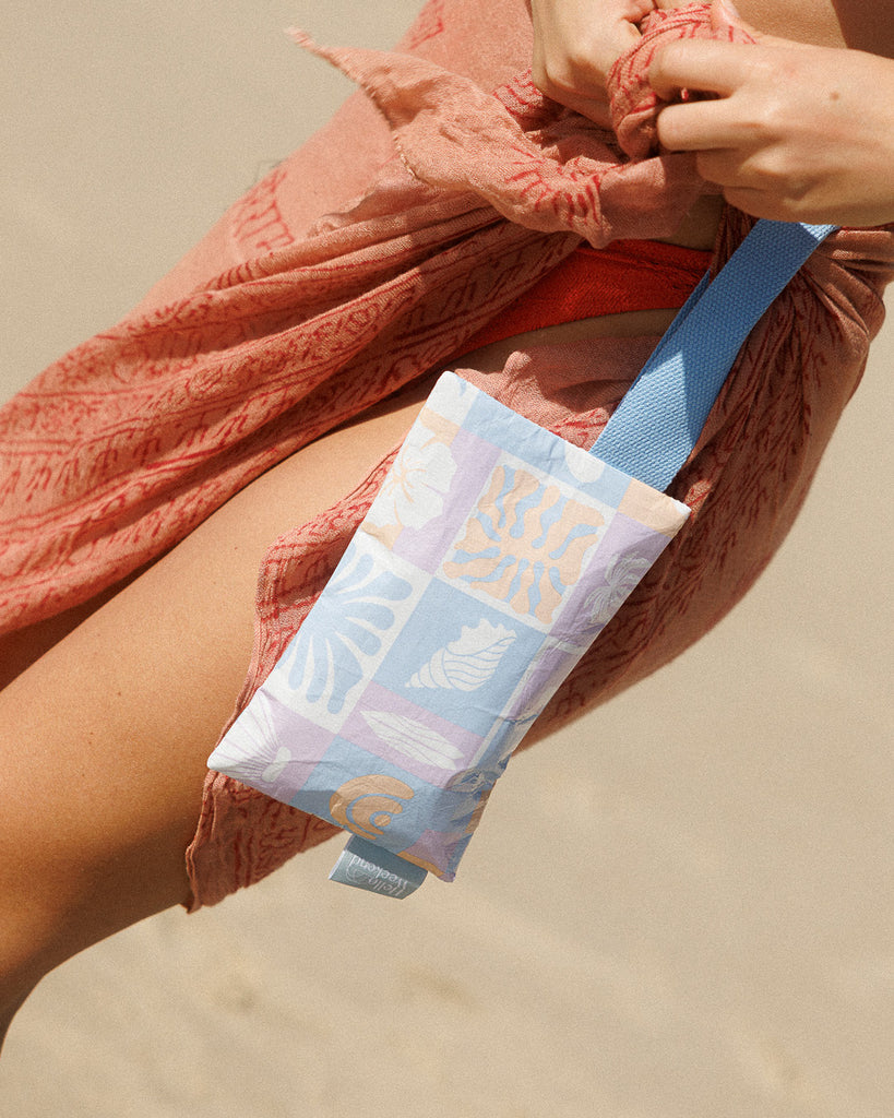 A small zippered pouch with a blue wrist strap, featuring a patchwork pattern of light blue, lavender, and peach squares with white floral and leaf motifs. A small fabric tag on the right reads Hello Weekend. Lizzie  Print - Phone Pouch - Reusable bags online | Daily bags | Shopper bags | Weekender bags  Hello Weekend
