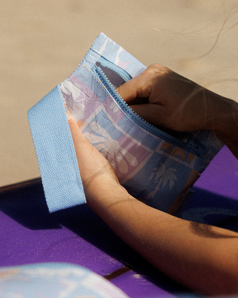 A small zippered pouch with a blue wrist strap, featuring a patchwork pattern of light blue, lavender, and peach squares with white floral and leaf motifs. A small fabric tag on the right reads Hello Weekend. Lizzie  Print - Phone Pouch - Reusable bags online | Daily bags | Shopper bags | Weekender bags  Hello Weekend