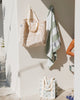 Tote bags and a towel hanging on a wall with a neutral background. Sandy Print - Weekender Bag - Reusable bags online | Daily bags | Shopper bags | Pouches  Hello Weekend Beige canvas handles | Beach Bags