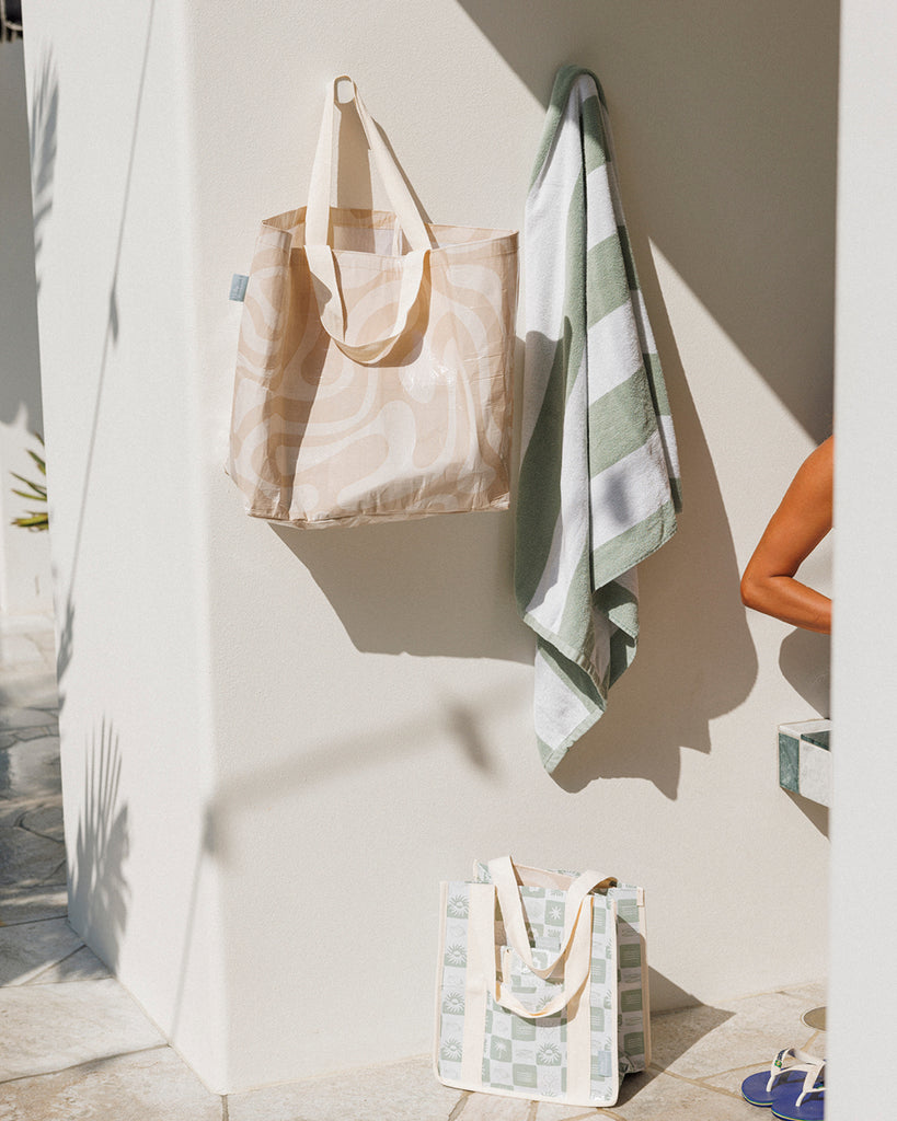 Tote bags and a towel hanging on a wall with a neutral background. Sandy Print - Weekender Bag - Reusable bags online | Daily bags | Shopper bags | Pouches  Hello Weekend Beige canvas handles | Beach Bags