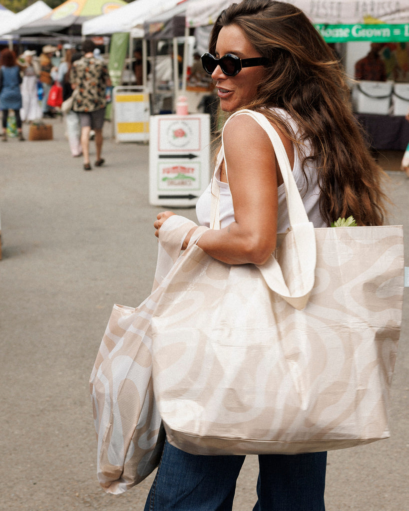 Woman carrying a large beige tote bag at an outdoor market. Sandy Print - Weekender Bag - Reusable bags online | Daily bags | Shopper bags | Pouches  Hello Weekend Beige canvas handles | Beach Bags